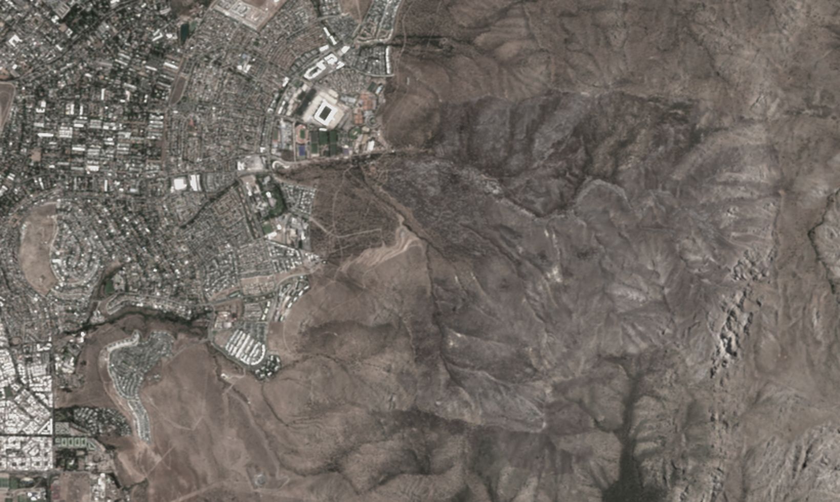 Imagen Sentinel-2 del 17 de enero de 2026 en tonos apagados, con los cerros al oriente de San Carlos de Apoquindo. Encima, en capas: la cicatriz del incendio en naranjo, el contorno blanco de la cuenca, las quebradas en celeste y la ruta de drenaje en amarillo desde la salida al borde urbano hacia el poniente.